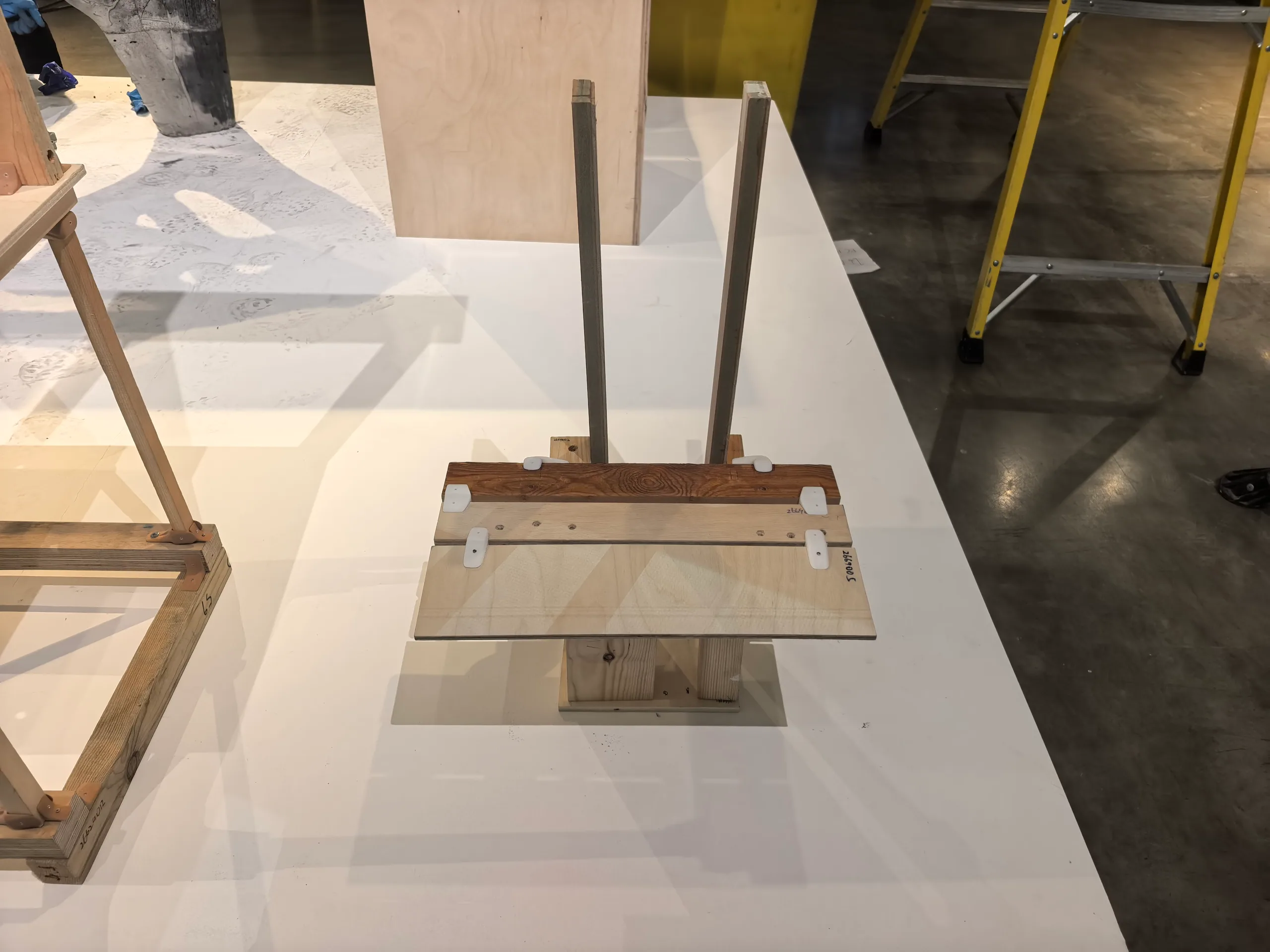 The Bartlett Autumn Show 2026: Khora 2: a chair of three boards on two timber blocks with two tall posts, joined with white connectors